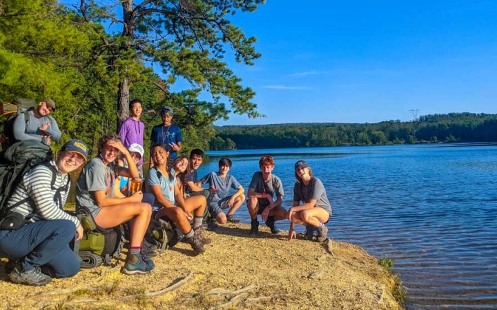 a group of teens sit next to a body of water on an outdoor expedition with outward bound